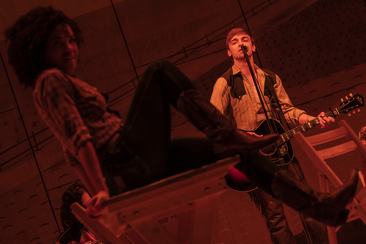 Production photo of Curly holding guitar in background, serenading a delighted-looking Laurey in foreground, all washed in red light