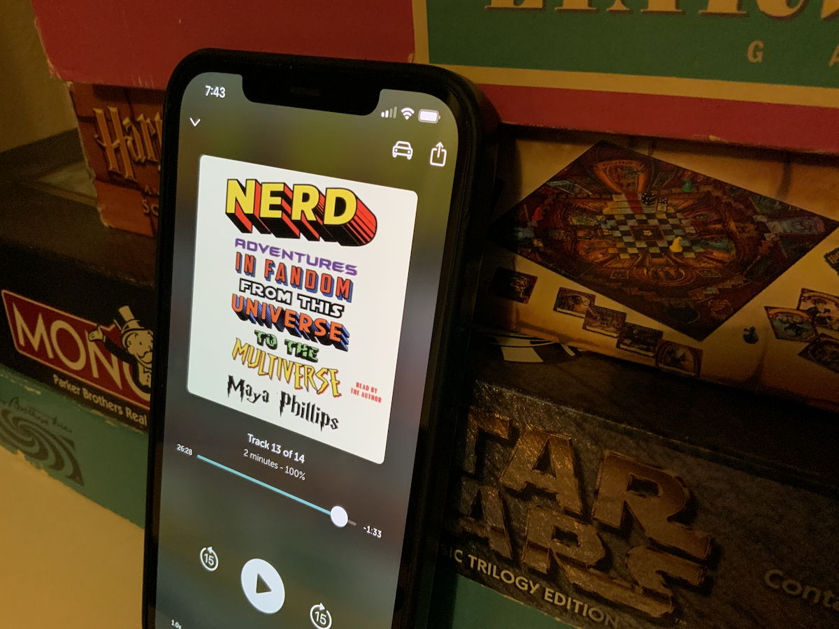 Photo of iPhone leaning against stack of board games including Harry Potter and Star Wars games. Phone is displaying audiobook player, featuring Maya Phillips's "Nerd: Adventures in Fandom from This Universe to the Multiverse."